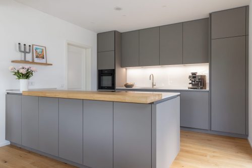 Grifflose Küche in der zeitlosen Farbe Fango. Gradlinig und ohne Schnickschnack wirkt diese Küche sehr clean und modern, aber nicht langweilig und durch die Barplatte aus Holz trotzdem so einladend, dass man sich gerne mit einem Getränk an die Bar setzt. 